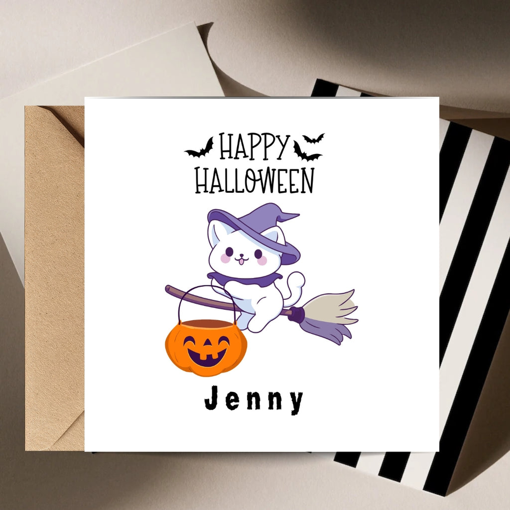 A square white greeting card with a Halloween design, shown with a kraft envelope behind it and a striped box at the side. The card reads 'Happy Halloween' above a cartoon white cat in a purple witch hat riding an orange jack-o'-lantern bucket broom, with the name 'Jenny' printed below.