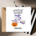 A square white greeting card with a Halloween design, shown with a kraft envelope behind it and a striped box at the side. The card reads 'Happy Halloween' above a cartoon white cat in a purple witch hat riding an orange jack-o'-lantern bucket broom, with the name 'Jenny' printed below.