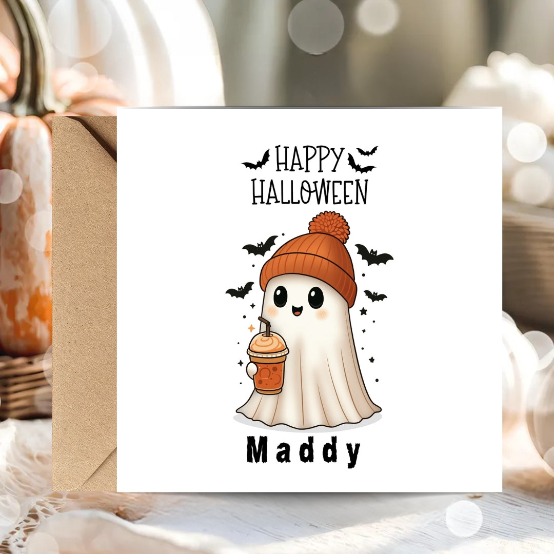 A white square Halloween greeting card with a cute ghost illustration wearing an orange beanie and holding an iced coffee, with the text 'HAPPY HALLOWEEN' above and 'Maddy' below. The card is shown standing in front of a kraft envelope, with blurred pumpkins and warm bokeh lights in the background.