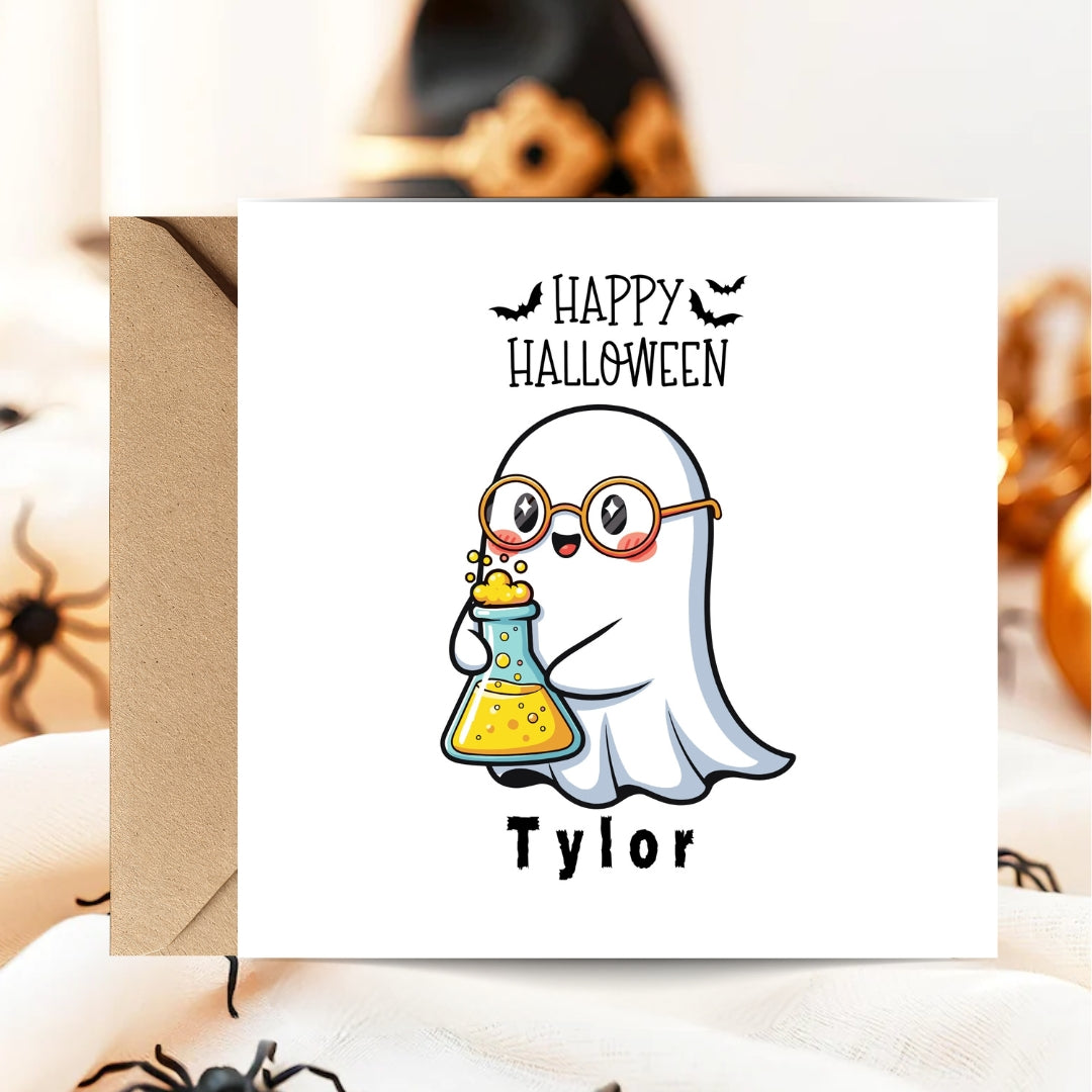 A square white Halloween greeting card with a kraft envelope, featuring a cartoon ghost wearing round glasses and holding a yellow flask under the words 'Happy Halloween' and above the name 'Tylor'. The card is photographed in a styled setting with blurred spider and pumpkin props.