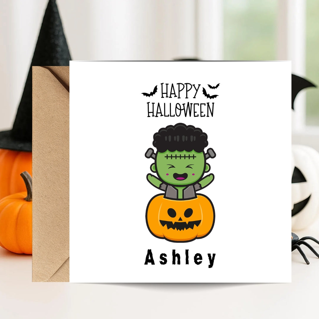 A square white Halloween greeting card with a kraft envelope, featuring a cartoon Frankenstein character sitting on a jack-o'-lantern and the text 'Happy Halloween' and 'Ashley'. The card is styled on a tabletop with pumpkins, a witch hat, and a spider in the background.