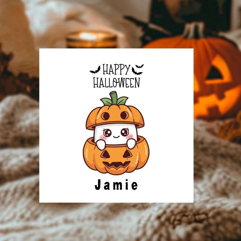 A square white card with a cute Halloween illustration, placed on a cozy blanket with a lit candle and a carved pumpkin in the background. The card reads 'Happy Halloween' with bats, a cartoon ghost inside a jack-o'-lantern, and the name 'Jamie' below.