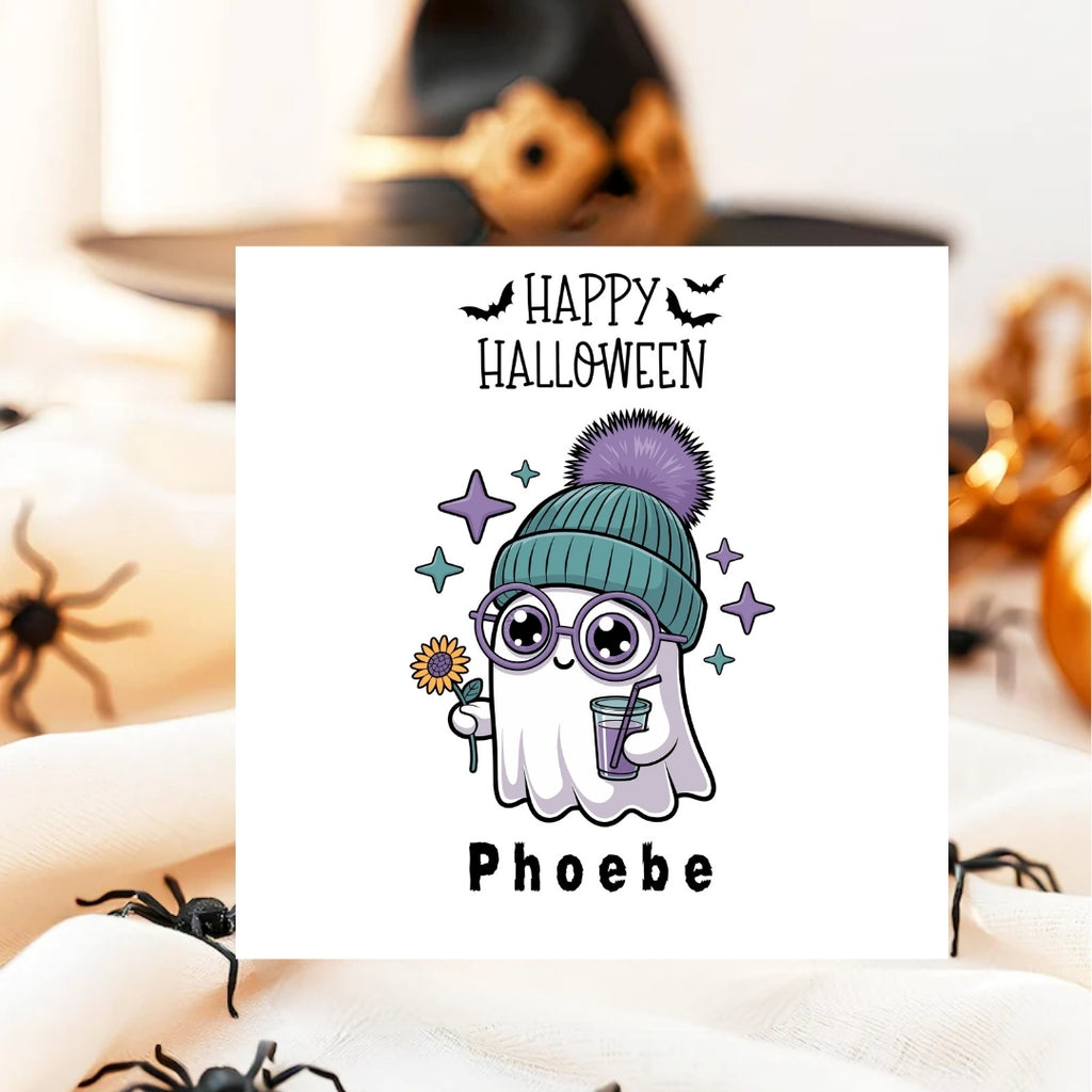 A square white Halloween greeting card featuring a cute ghost illustration wearing a teal beanie with a purple pom-pom, glasses, and holding a flower and drink. The card reads 'Happy Halloween' at the top and 'Phoebe' at the bottom.