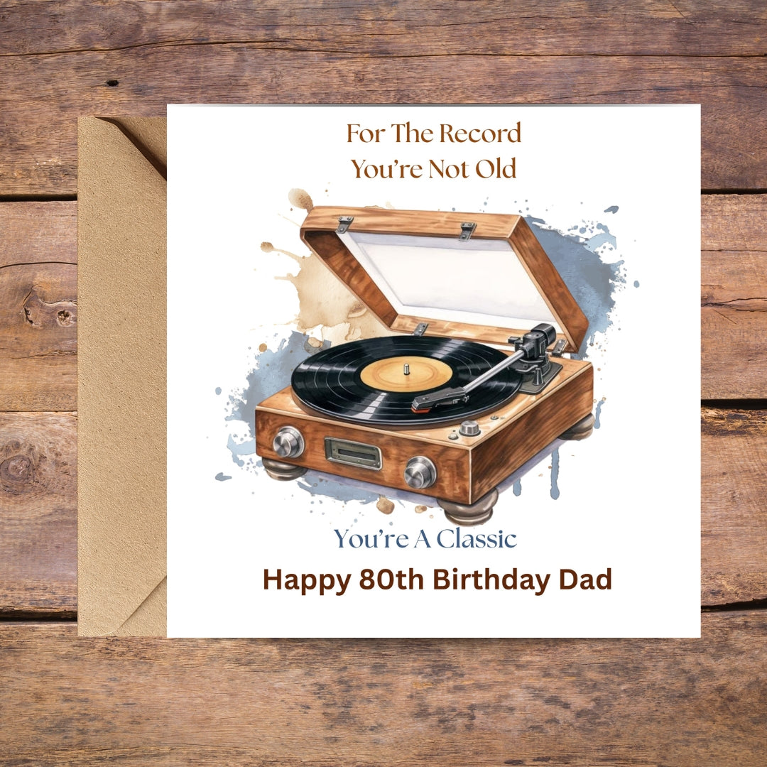 A square birthday card with a kraft envelope, displayed on a rustic wooden background. The card features a watercolor illustration of a wooden turntable with a vinyl record.