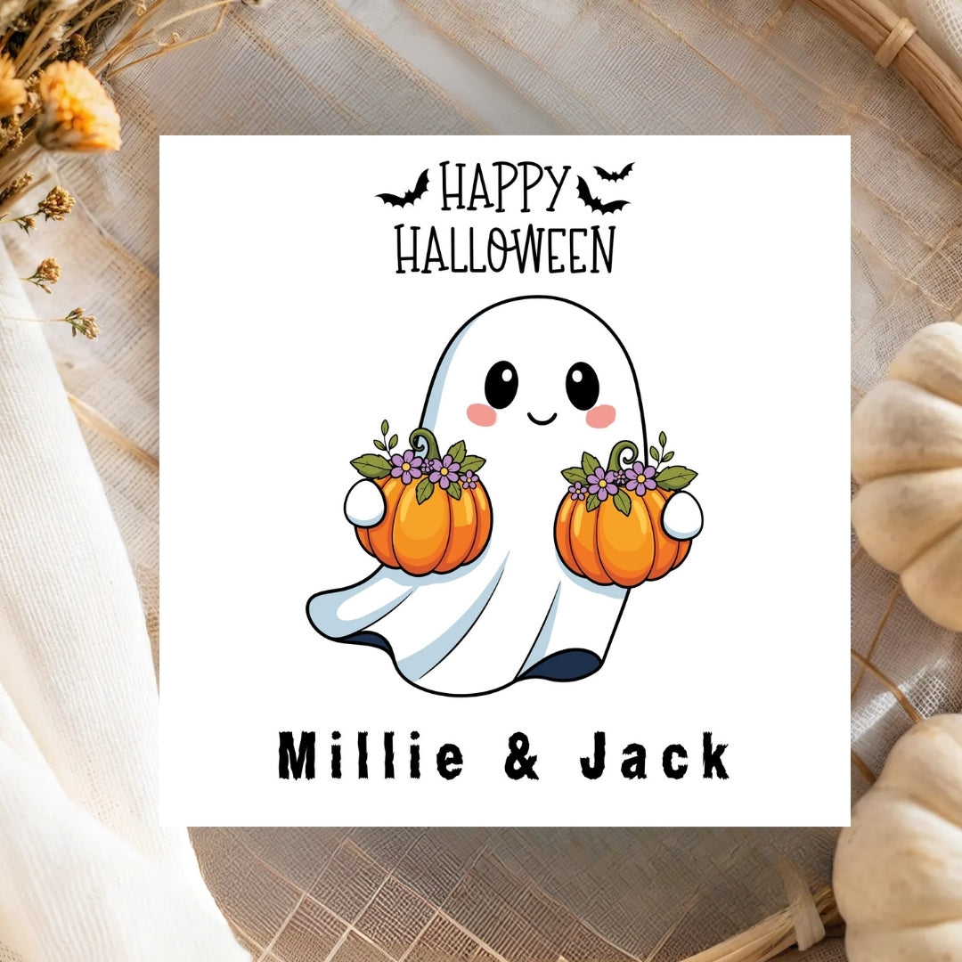 A square white Halloween greeting card or print featuring a cute cartoon ghost holding two orange pumpkins with purple flowers, with the text 'HAPPY HALLOWEEN' above and 'Millie & Jack' below. The card is styled on a woven mat with dried flowers and pumpkins around it.