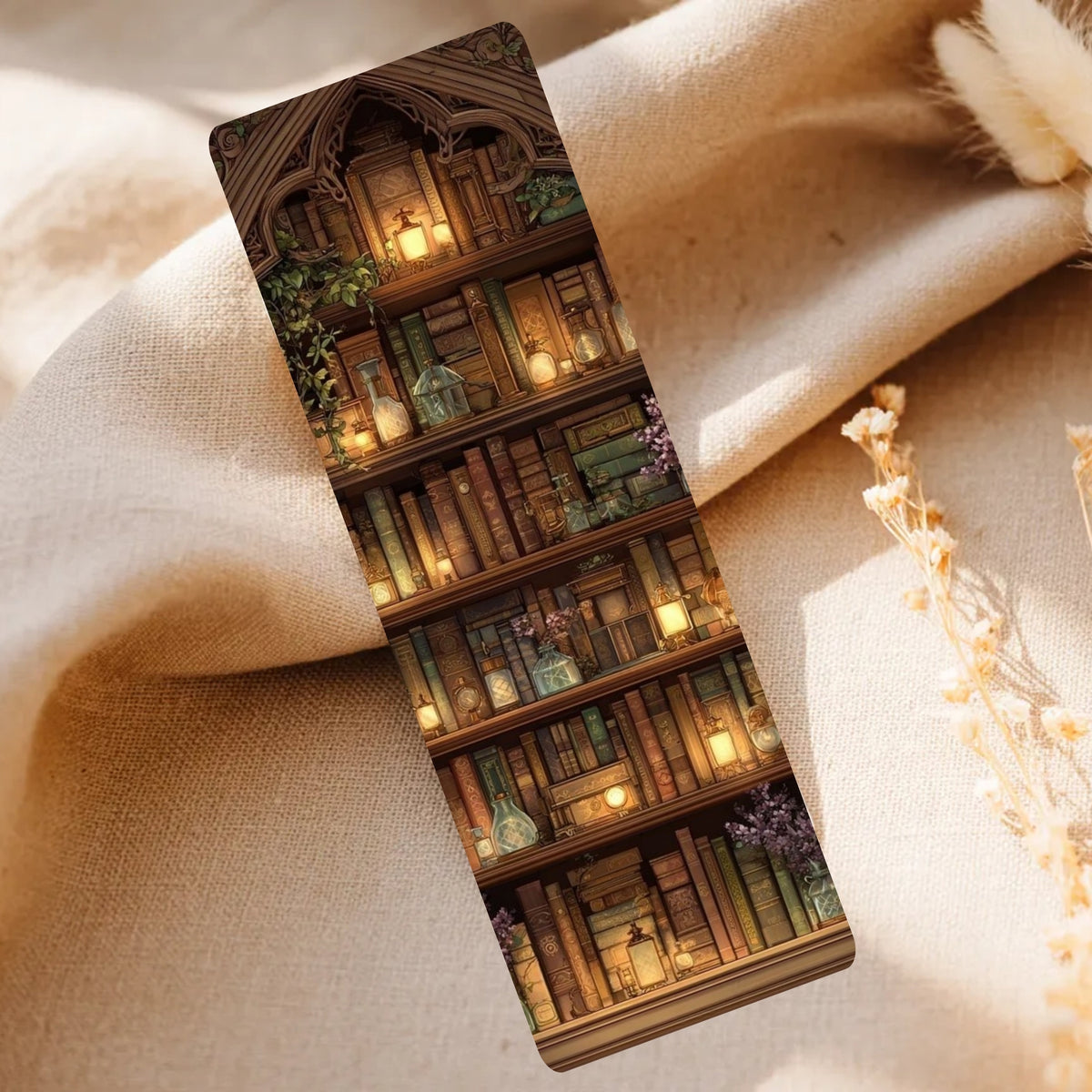 A slim rectangular bookmark or card featuring a printed illustration of a cozy library bookshelf with candles, books, and greenery, placed on draped beige fabric with dried flowers nearby.