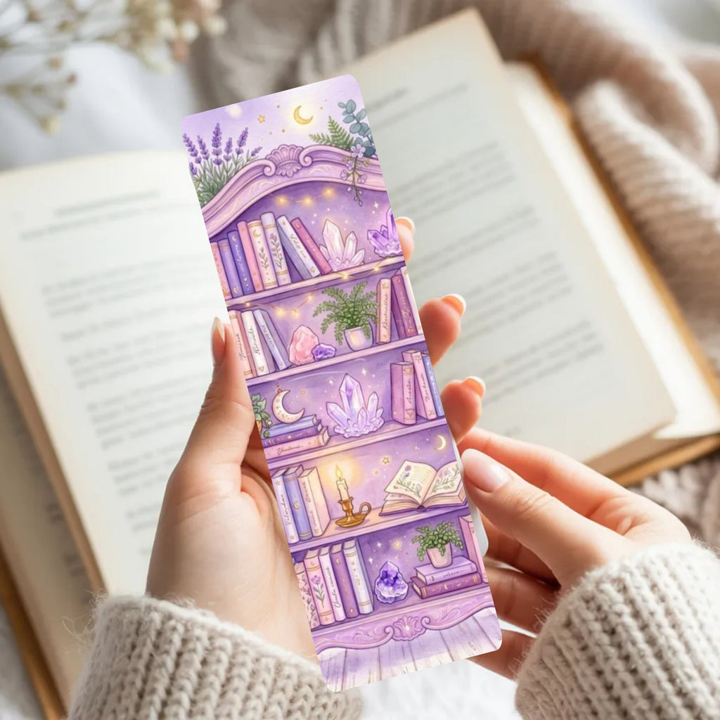 A person holds a lavender bookmark featuring an illustrated bookshelf with crystals, plants, and stars, set against an open book and soft knit sweater.