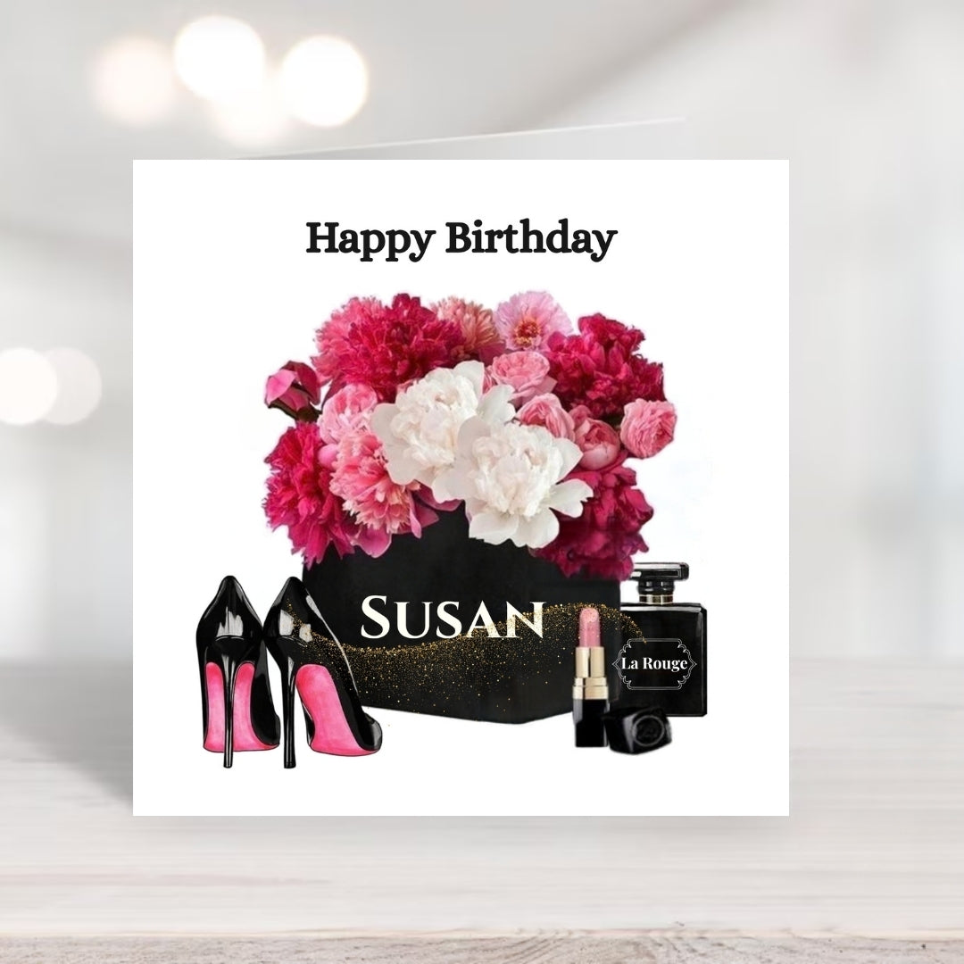 A square birthday greeting card displayed on a light wood surface with a soft blurred background. The card features the text 'Happy Birthday' above a floral arrangement in a black pot labeled 'SUSAN', flanked by black high-heeled shoes with pink soles, a lipstick tube, and a perfume bottle labeled 'La Royale'.