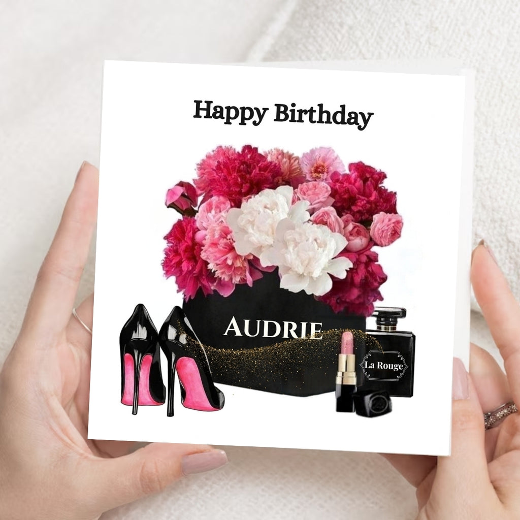 A hand-held square birthday card with the text 'Happy Birthday' above an illustration of pink and white flowers in a black box labeled 'AUDRIE,' along with black high heels with pink soles, a pink lipstick tube, and a black perfume bottle labeled 'La Rouge.' Two hands are holding the card against a soft light background.