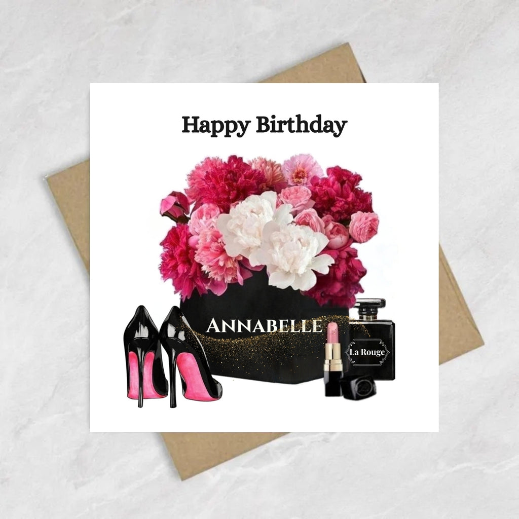 A square birthday card with a white background featuring the text 'Happy Birthday' above a floral bouquet, black high heels, a black box labeled 'ANNABELLE', and a black perfume bottle labeled 'La Royale'. The card is placed on top of brown envelopes on a light marble-like surface.
