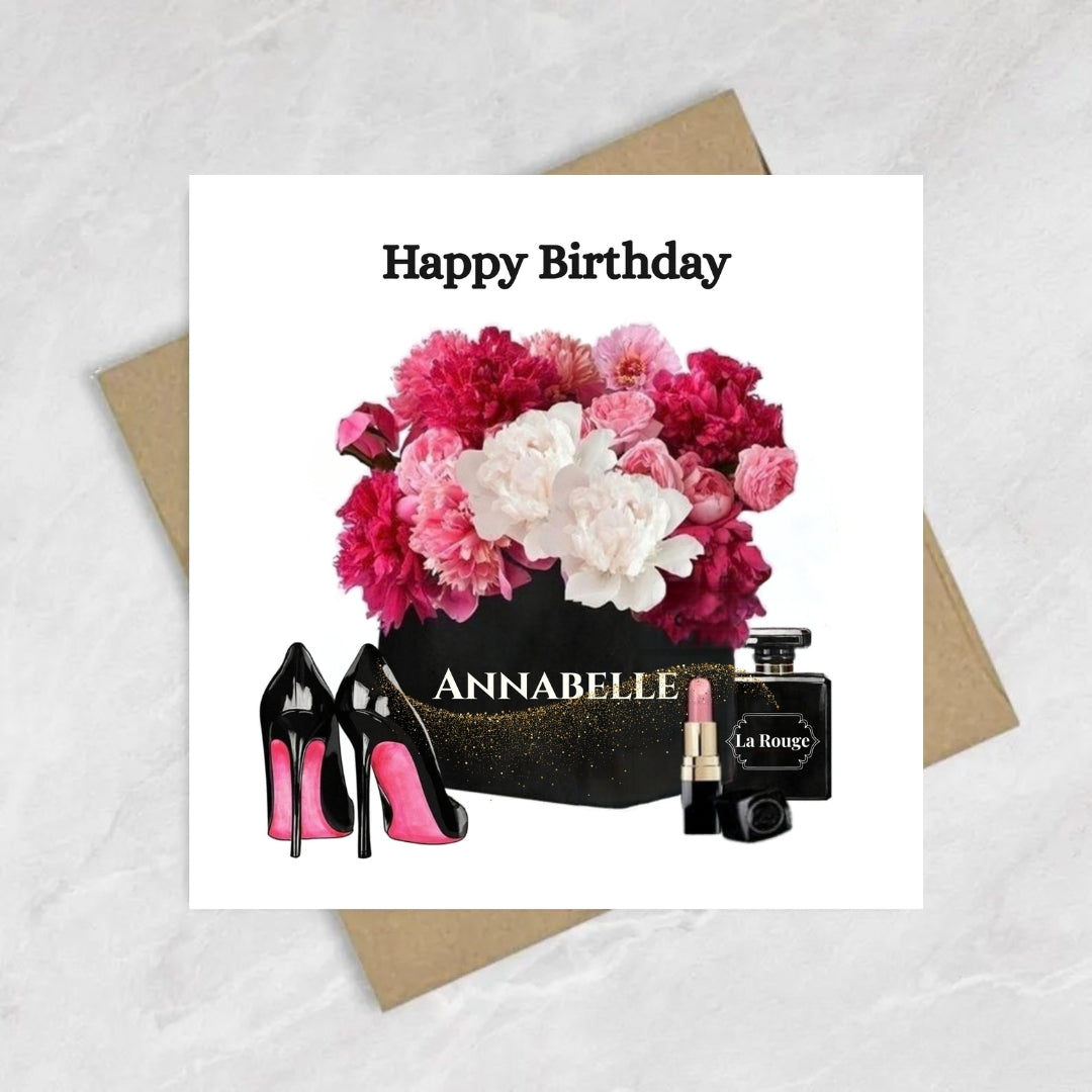 A square birthday card with a white background featuring the text 'Happy Birthday' above a floral bouquet, black high heels, a black box labeled 'ANNABELLE', and a black perfume bottle labeled 'La Royale'. The card is placed on top of brown envelopes on a light marble-like surface.