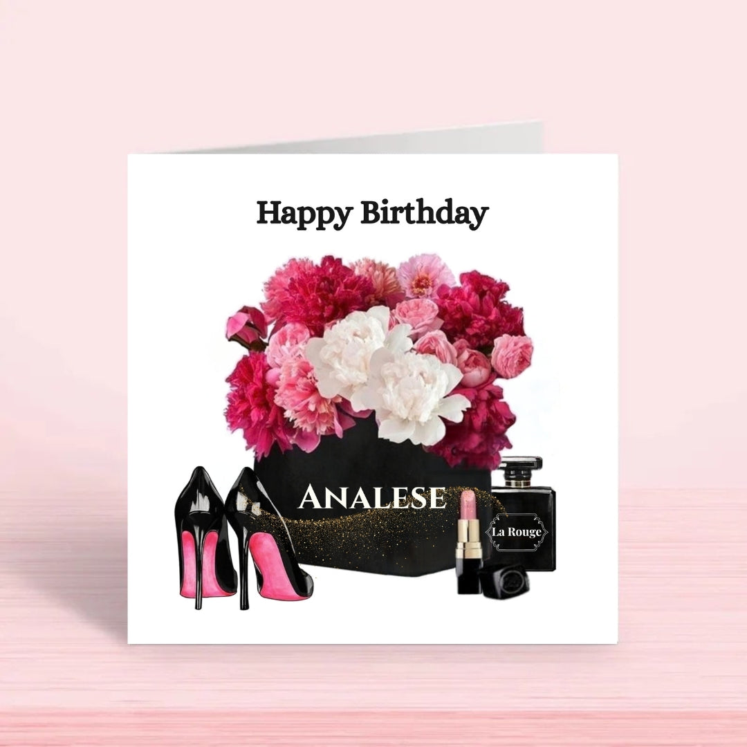A square white greeting card with a floral and fashion-themed birthday design, shown standing on a pale pink surface. The card reads 'Happy Birthday' above a bouquet of pink and white flowers, black high heels with bright pink soles, a black box labeled 'ANALESE', and a black perfume bottle labeled 'La Rouge'.
