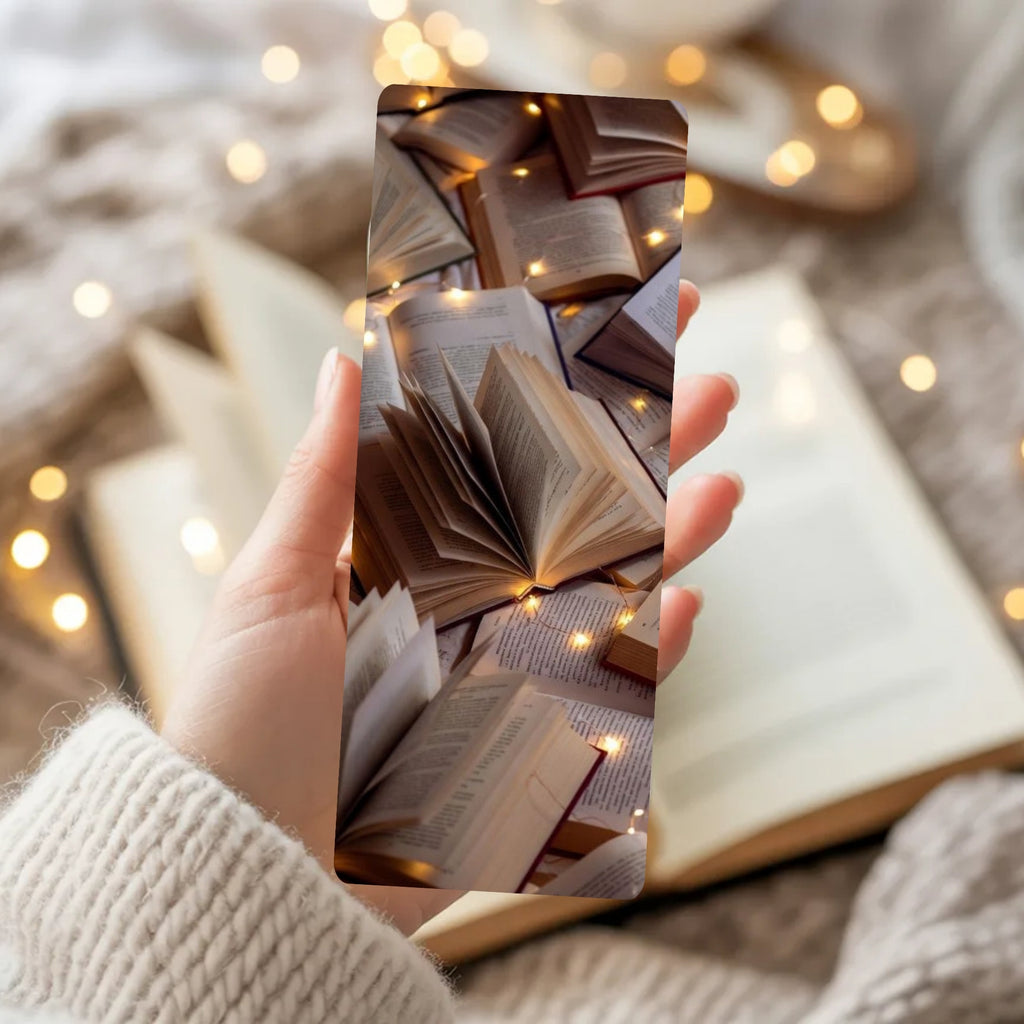 A hand in a cream knit sweater holds a rectangular phone case featuring an artistic print of open vintage books and warm fairy lights. The background is a cozy, softly blurred setting with more books and warm bokeh lights.