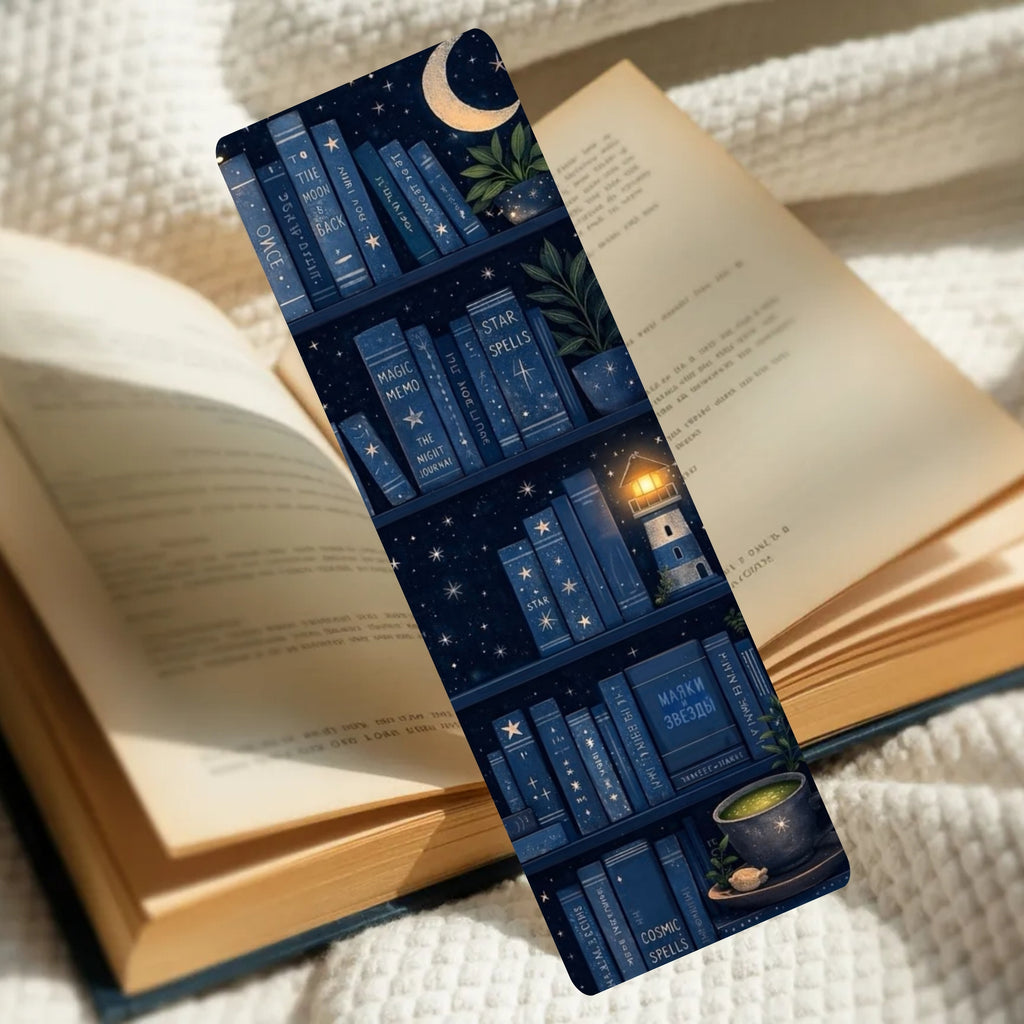 A long rectangular bookmark or book divider printed with a navy blue bookshelf scene, stars, moon, plants, and a glowing lighthouse, placed across an open book on a cream knit blanket.