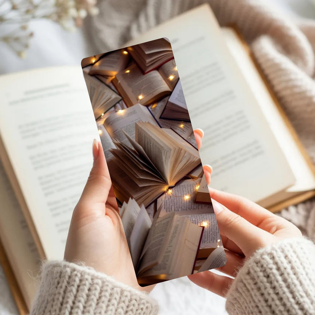 A person holds a slim rectangular book bookmark or card featuring a printed design of stacked open books with warm fairy lights, in front of an open book and cozy knit sweater.