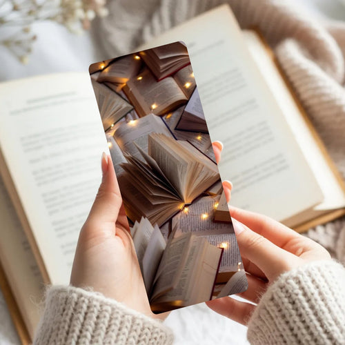 A person holds a slim rectangular book bookmark or card featuring a printed design of stacked open books with warm fairy lights, in front of an open book and cozy knit sweater.