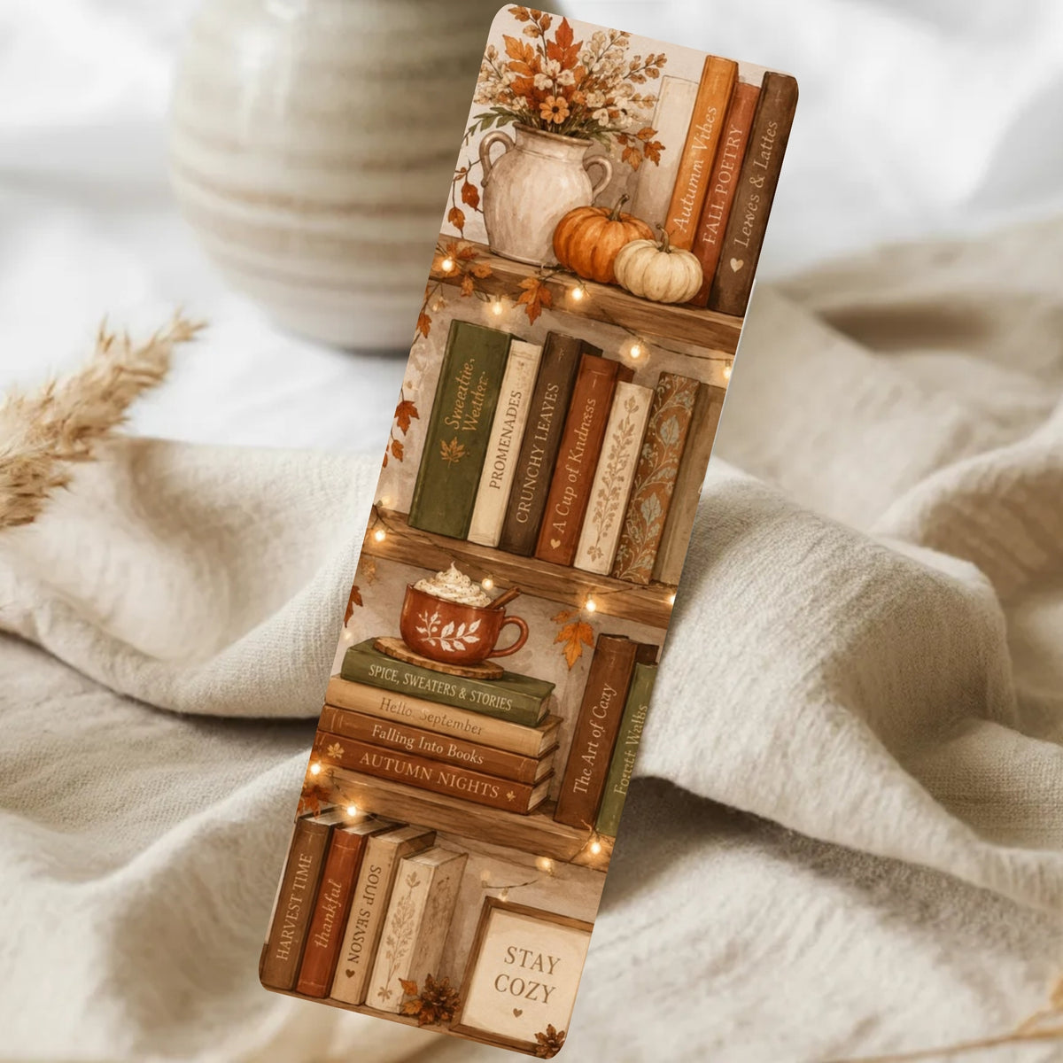 A slim rectangular phone case with a cozy autumn-themed bookshelf print, shown resting on draped cream fabric with a soft blurred background. The design features vintage books, small pumpkins, a mug of hot cocoa with marshmallows, warm string lights, and fall leaves.