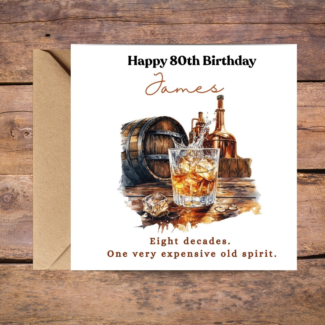 A personalized 80th birthday greeting card with an envelope, displayed on a rustic wooden background. The card features a watercolor illustration of a whiskey glass, a barrel, and bottles, with the text 'Happy 80th Birthday James' and 'Eight decades.