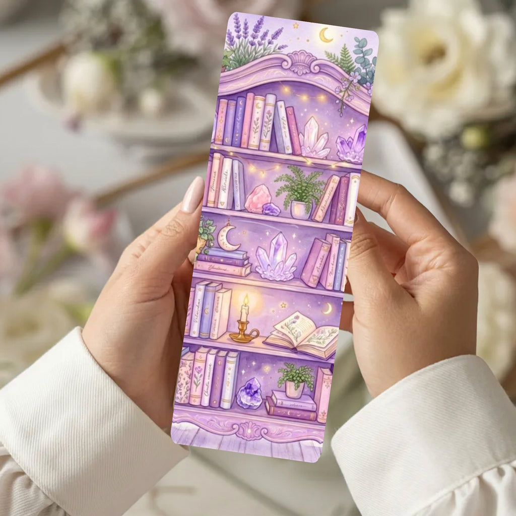 A person holds a long rectangular bookmark or card featuring a whimsical purple bookshelf illustration with crystals, books, plants, and moon motifs. The background is softly blurred with flowers and neutral decor.