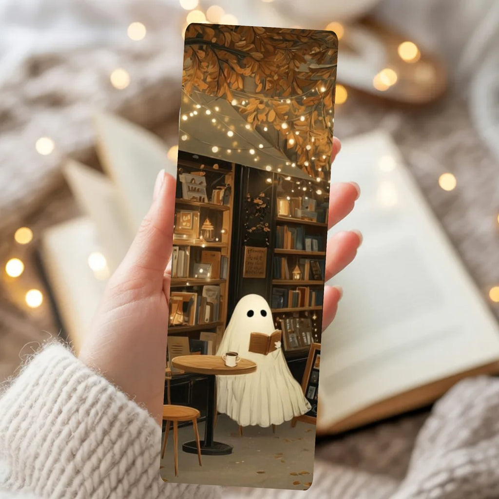 A hand in a cream knit sweater holds a slim rectangular printed card or bookmark featuring an illustrated cozy library scene with a ghost-like figure reading under warm string lights. The background is softly blurred with open books and warm bokeh lights.