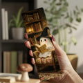 A hand holds a slim rectangular printed item, likely a bookmark or small card, featuring a cute ghost character in a witch hat reading in a cozy armchair by a bookshelf. The background is a softly blurred indoor scene with plants and books.