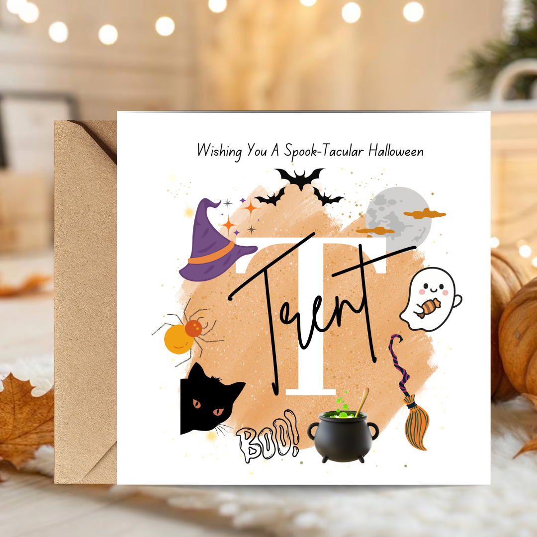 A square Halloween greeting card with a kraft envelope, displayed on a cozy tabletop with warm bokeh lights and autumn decor in the background. The card features a white background with a large decorative letter 'T' over a peach watercolor splash, surrounded by cute illustrations of a witch's hat, spider, cat, cauldron, broom, moon, bats, and a ghost.