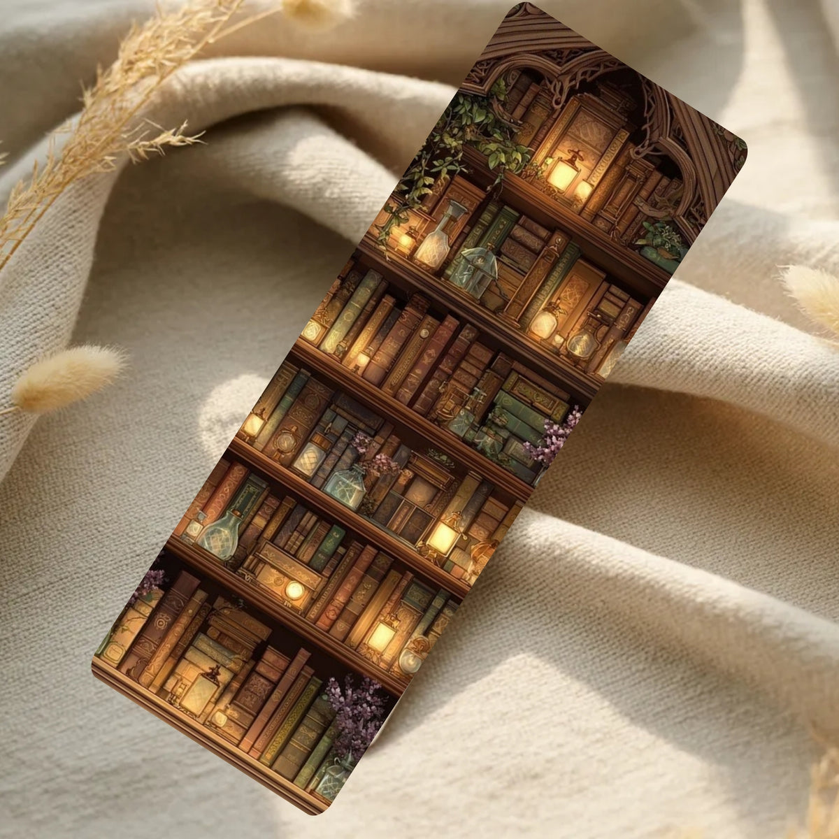 A rectangular bookmark featuring an illustrated library bookshelf scene with glowing candles and books, placed on draped cream fabric with dried grass accents.