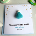 A personalized new baby welcome card featuring a small turquoise knitted baby hat with a white pom-pom on a white card with gold stars and the text 'Welcome To The World Baby Jackson 16.10.