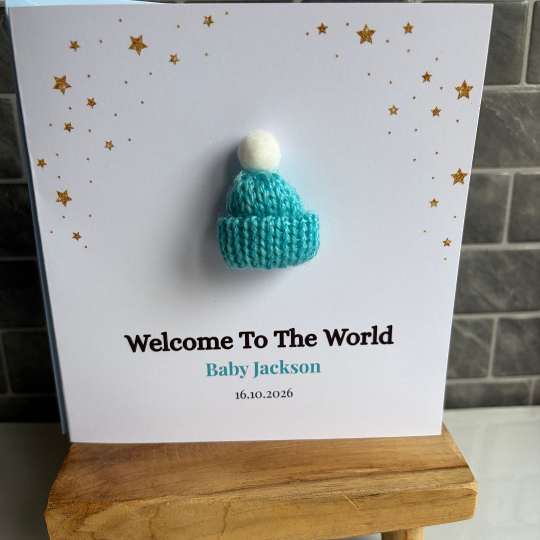 A personalized baby welcome card propped on a wooden stand, featuring a small knitted teal beanie with a white pom-pom attached to the center. The card is printed with gold stars, the text 'Welcome To The World', 'Baby Jackson', and the date '16.