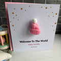 A personalized baby welcome card with a pink knitted beanie charm attached to the front. The card is white with gold star details, the text 'Welcome To The World Baby Amelia 16.