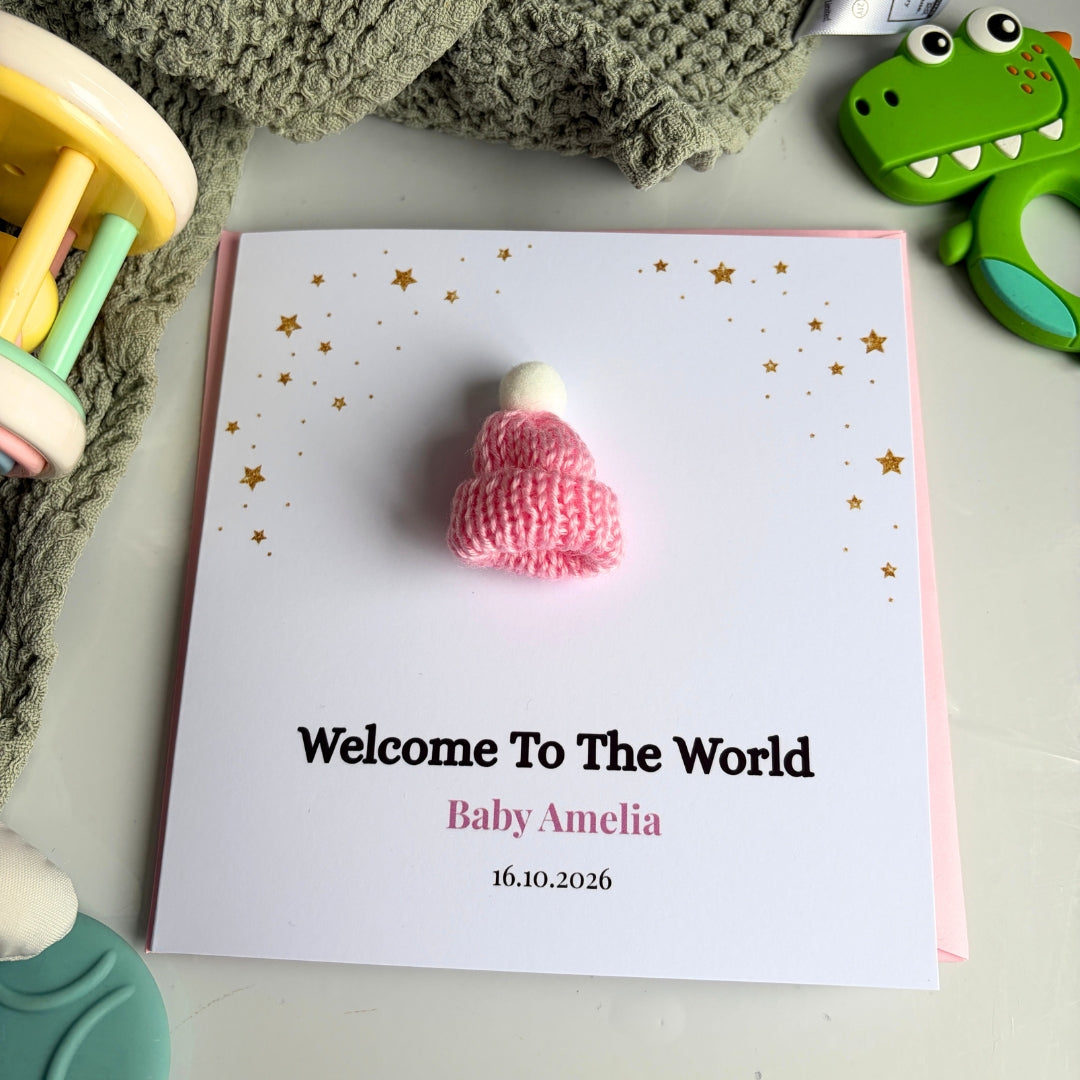 A personalized baby shower card with a pink knitted baby hat on a white card that reads 'Welcome To The World Baby Amelia 16.10.