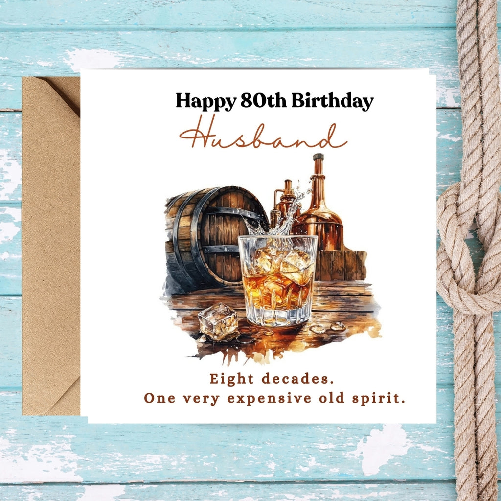 A square birthday card for an 80th birthday husband, displayed with a brown envelope on a distressed light blue wooden background next to a rope. The card features a watercolor illustration of an whiskey glass, bottles, and a barrel, with the text 'Happy 80th Birthday Husband' and 'Eight decades.