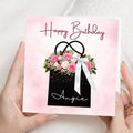 A person holds a square birthday card with a pink watercolor background, a black gift bag illustration with pink flowers, green leaves, gold confetti, and a white ribbon. The card reads 'Happy Birthday Angie' in script text.