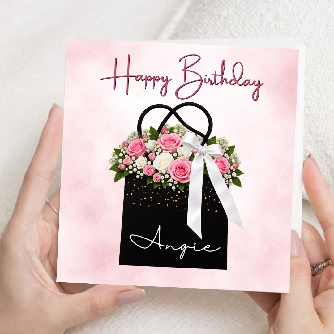 A person holds a square birthday card with a pink watercolor background, a black gift bag illustration with pink flowers, green leaves, gold confetti, and a white ribbon. The card reads 'Happy Birthday Angie' in script text.