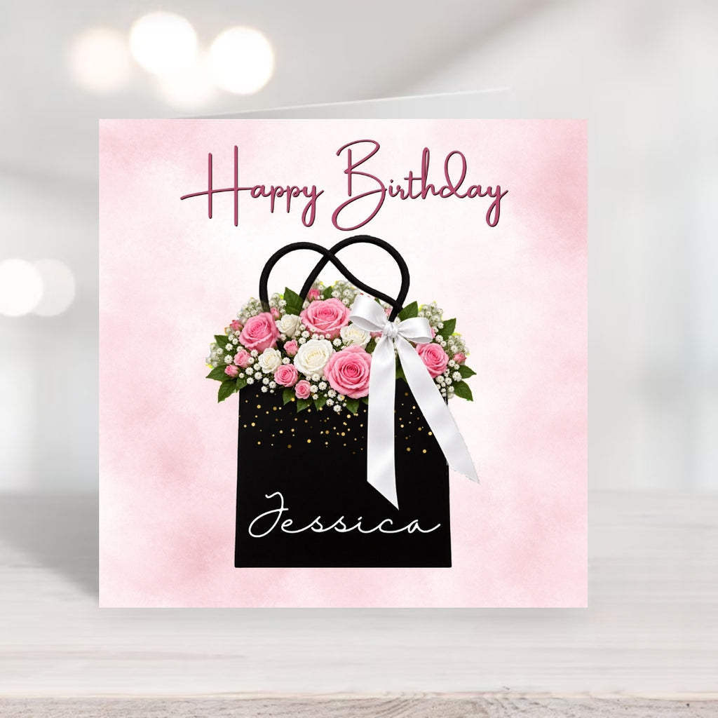 A square birthday card with a pink watercolor background, featuring a black gift bag illustration filled with pink and white flowers, a white ribbon, and the words 'Happy Birthday' and 'Jessica'.