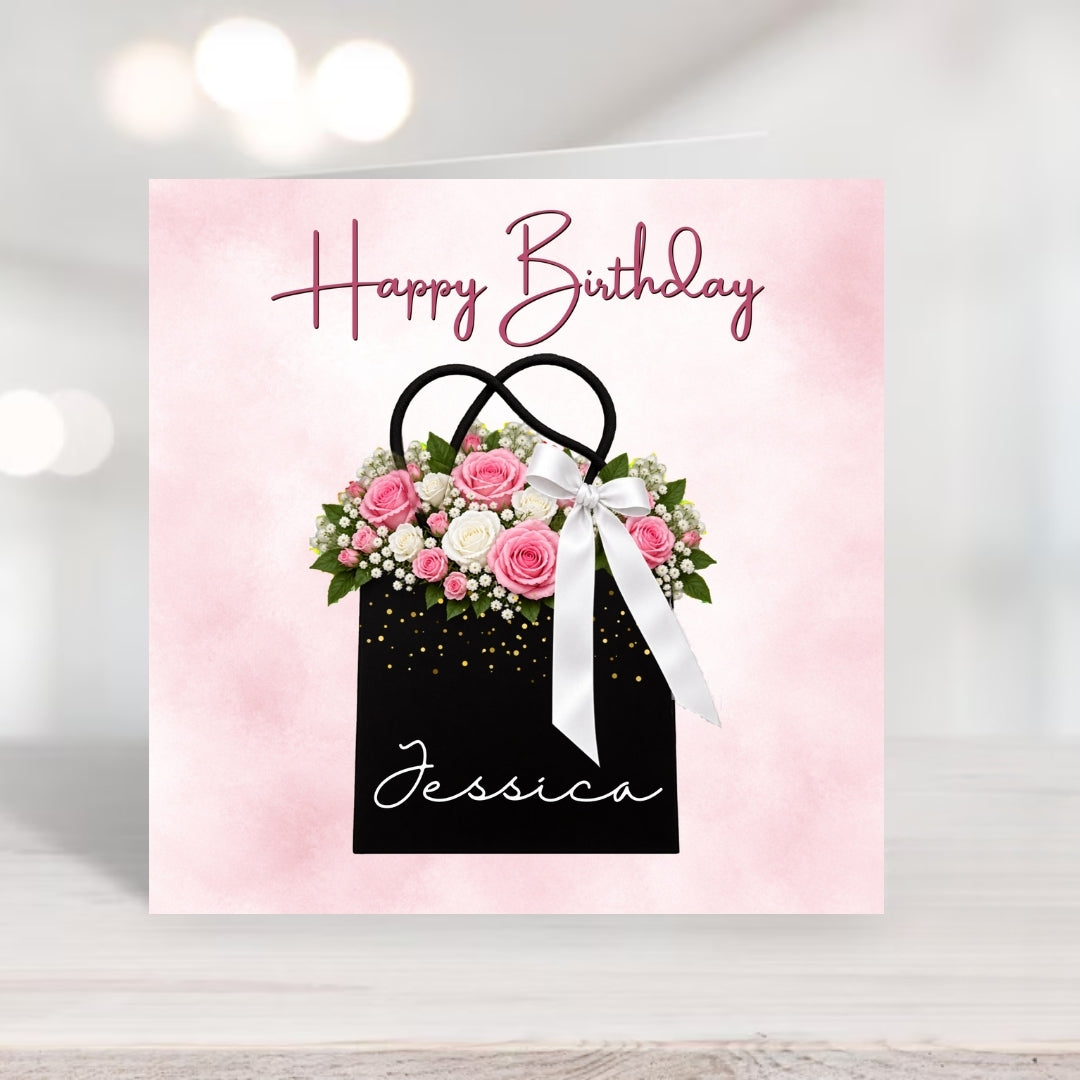 A square birthday card with a pink watercolor background, featuring a black gift bag illustration filled with pink and white flowers, a white ribbon, and the words 'Happy Birthday' and 'Jessica'.