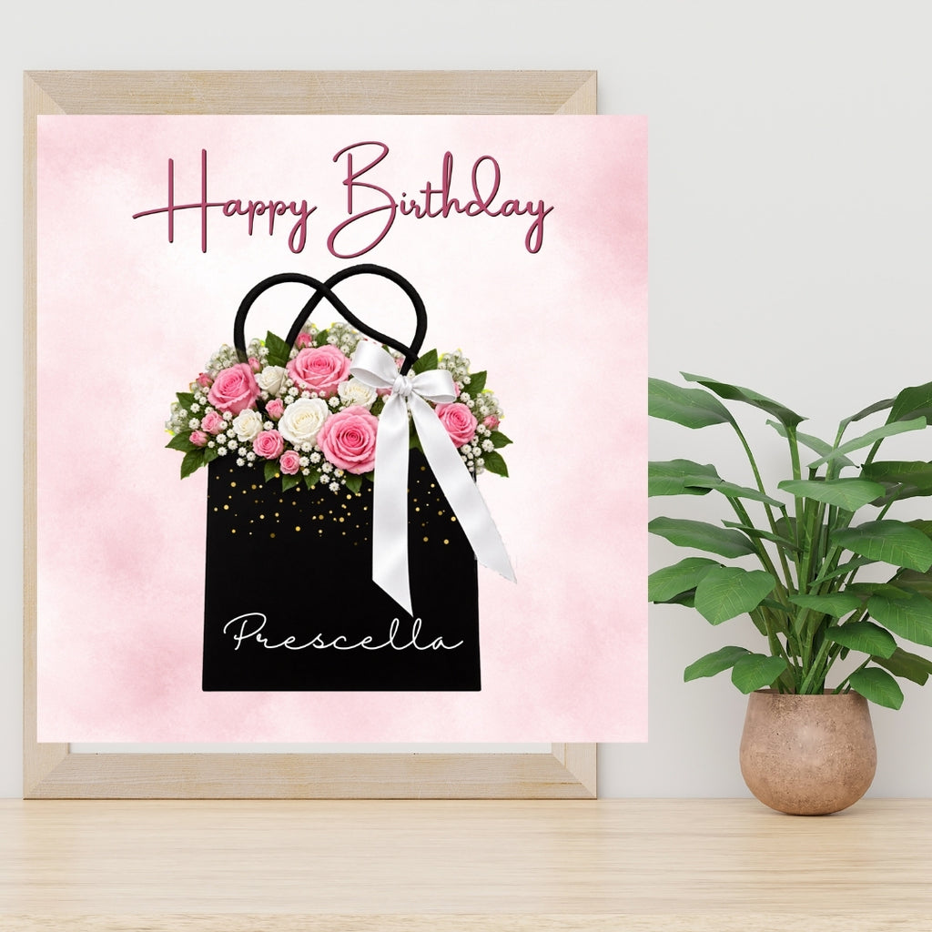 A framed personalized birthday card featuring a pink watercolor background, the words 'Happy Birthday' in script, and a black gift bag filled with pink and white roses. The card is displayed on a light wood surface next to a potted green plant.