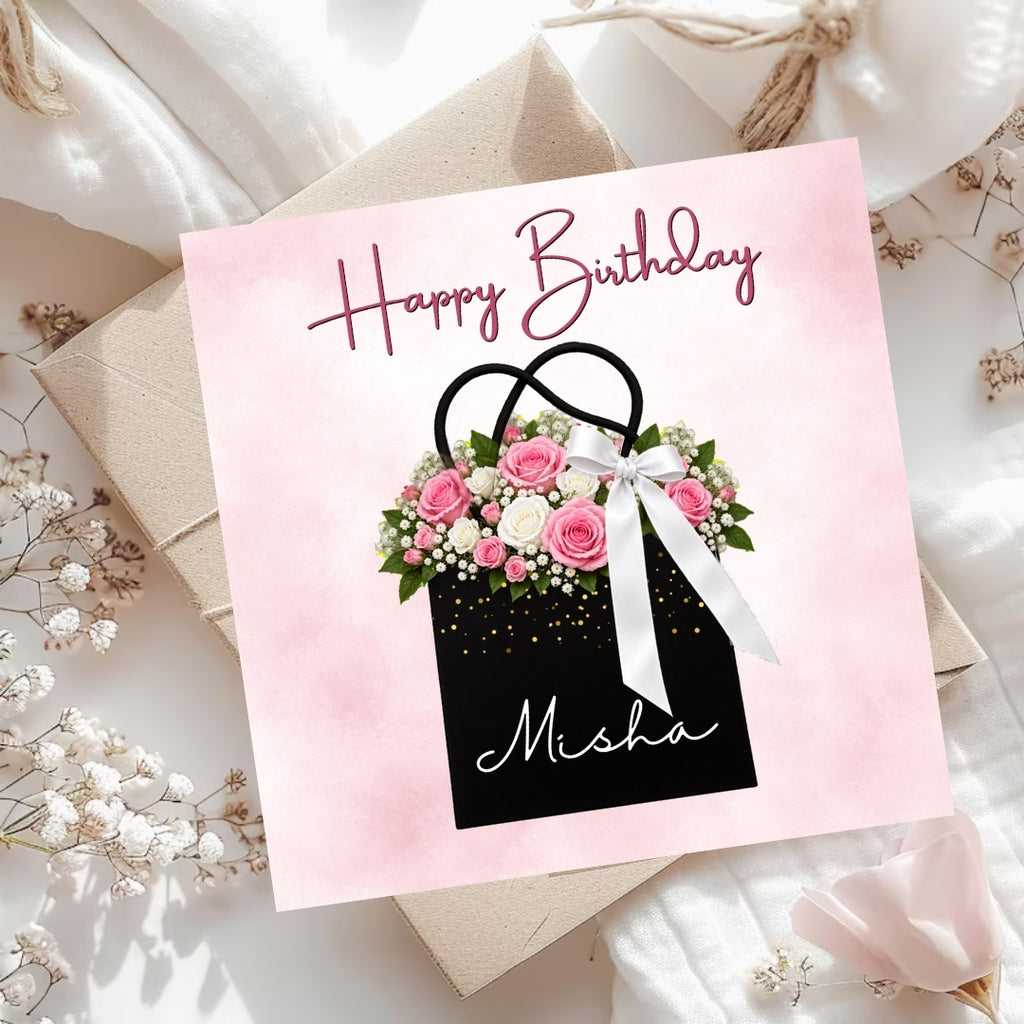 A square birthday card with a pink watercolor background, featuring a black gift bag illustration decorated with pink and white flowers, green leaves, and a white ribbon. The card reads 'Happy Birthday' in pink script at the top and 'Misha' in white script near the bottom.