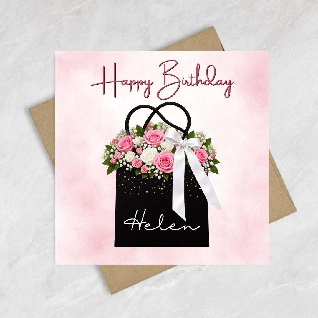 A square birthday card with a pink watercolor background, featuring a black gift bag filled with pink and white flowers, a white ribbon, and the name 'Helen' in white script. The card reads 'Happy Birthday' in pink cursive at the top.
