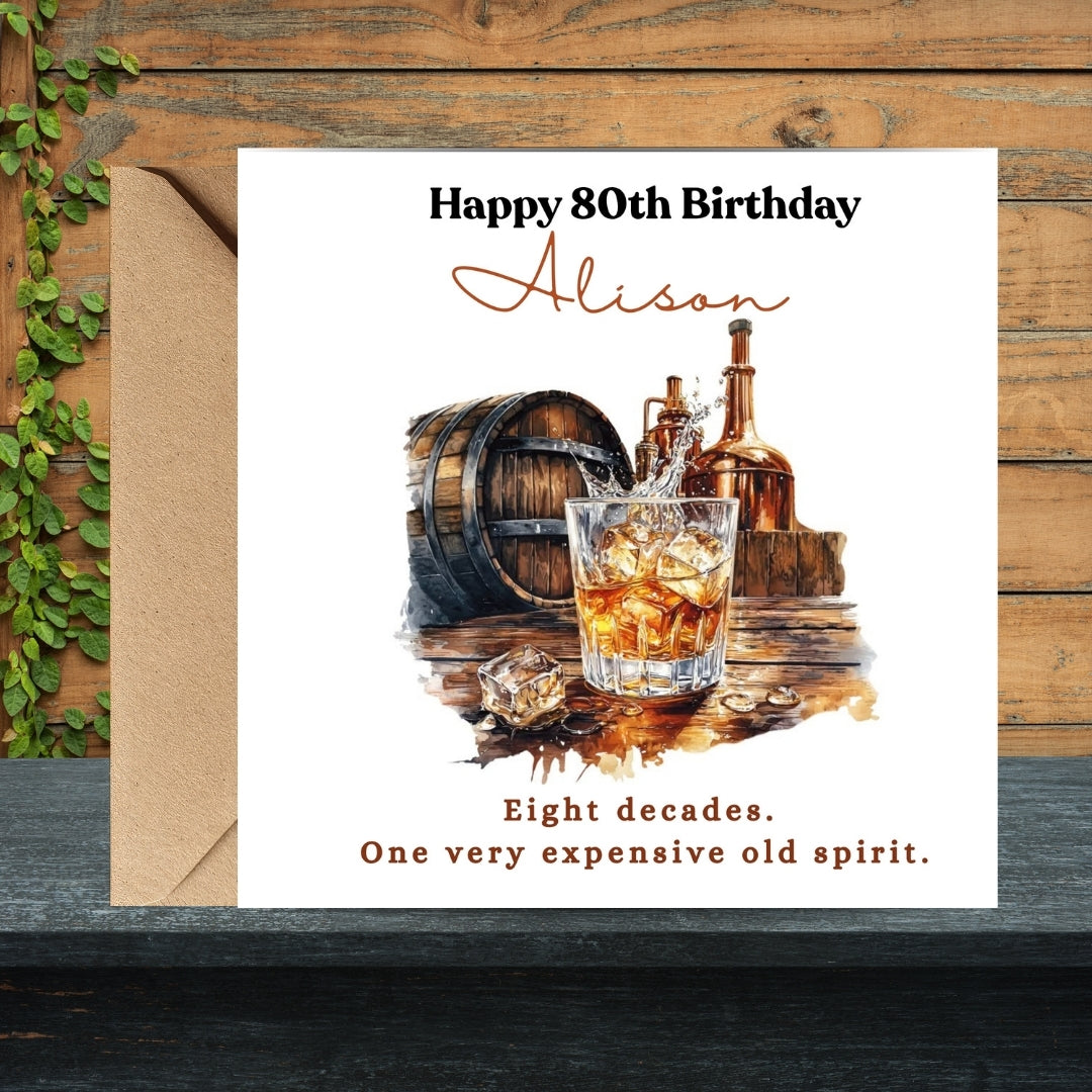 A square birthday card with a kraft envelope, displayed on a dark wooden surface next to green ivy. The card features a watercolor illustration of an whiskey glass, a barrel, and bottles, with the text 'Happy 80th Birthday Alison' and 'Eight decades.