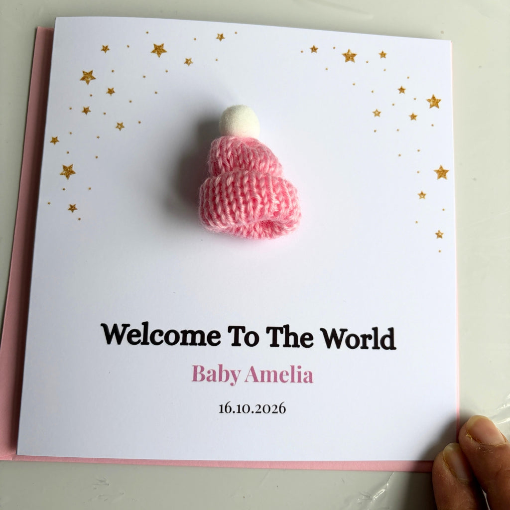 A personalized baby welcome card held by a hand, featuring a small pink and white knitted hat on a white card with gold stars and the text 'Welcome To The World Baby Amelia 16.10.