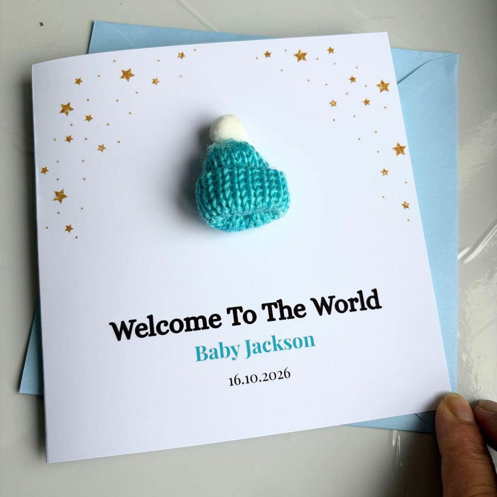 A personalized new baby card featuring a small turquoise knitted baby hat with a white pom-pom on a white card with gold stars and the text 'Welcome To The World Baby Jackson 16.10.