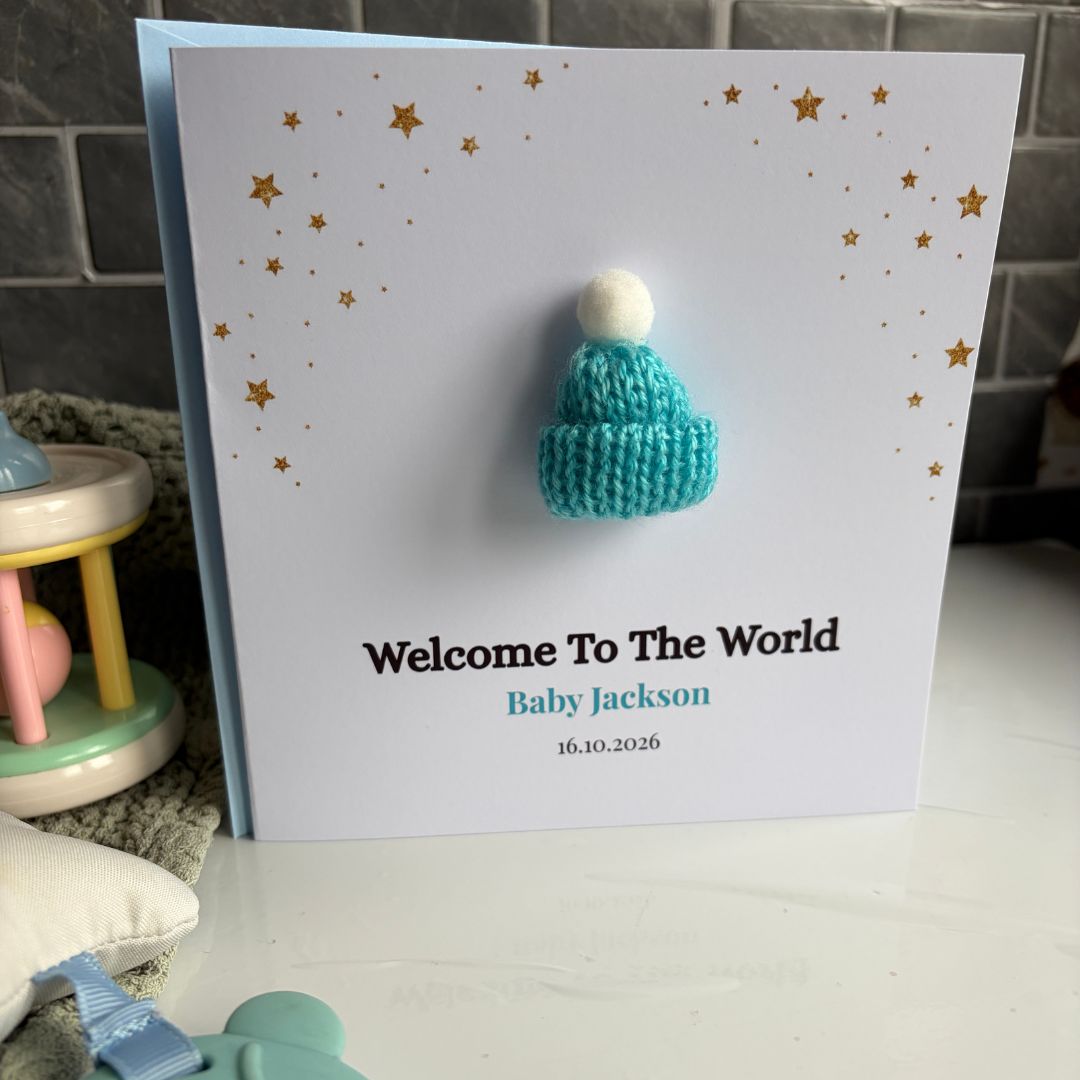 A personalized baby birth announcement card with a white background, gold star details, and a small knitted teal beanie with a white pom-pom attached to the front. The card is printed with the text 'Welcome To The World', 'Baby Jackson', and the date '16.