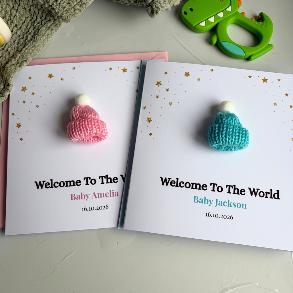 Two personalized baby welcome cards laid flat on a light surface, each featuring a small knitted baby hat in pink and teal with gold star accents. The left card reads 'Welcome To The V Baby Amelia 16.