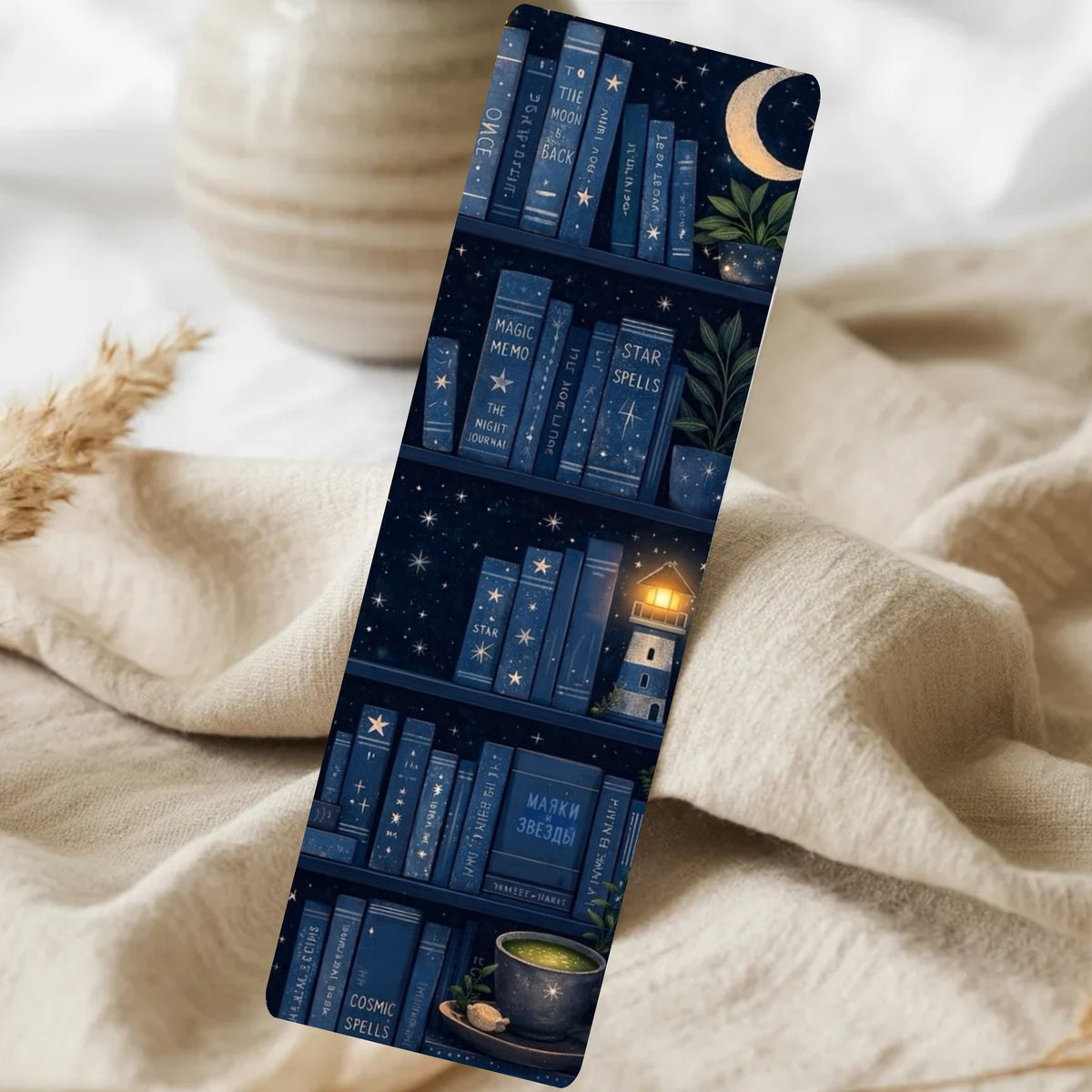 A slim rectangular bookmark or card featuring a dark navy bookshelf illustration with books, stars, a crescent moon, a small glowing lighthouse, and cozy props, shown resting on beige fabric with a blurred ceramic vase in the background.