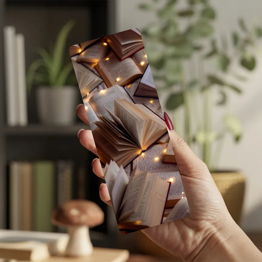 A hand holding a slim rectangular card or case printed with a collage of open books and warm fairy lights, shown in a softly blurred indoor setting with plants and shelves in the background.