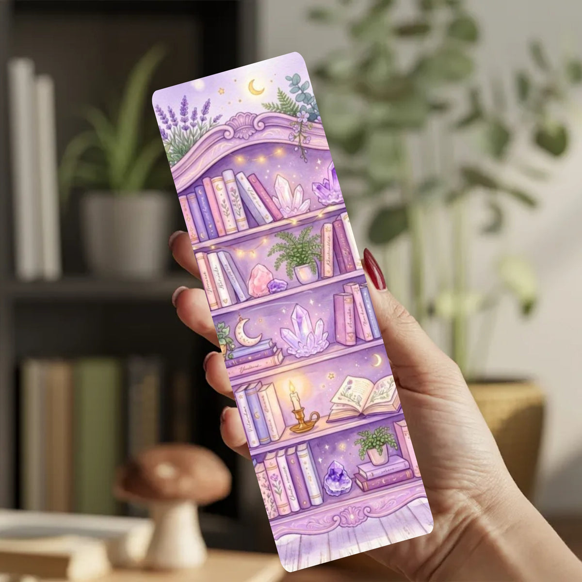 A hand holds a long, narrow decorative bookmark or card featuring a whimsical purple bookshelf illustration with books, crystals, plants, and moons, set against a softly blurred indoor background.