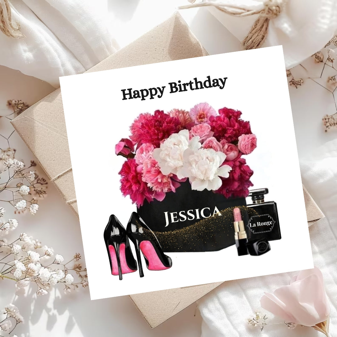 A square birthday card with a white background, featuring a graphic of a pink and white flower bouquet, black high heels with pink soles, a black perfume bottle labeled 'La Honey', and the name 'JESSICA' in white text. The card is styled on top of a kraft paper envelope and white fabric with dried flowers.